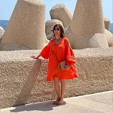 Een persoon in een oranje jurk staat op een betonnen oppervlak met grote stenen structuren op de achtergrond.