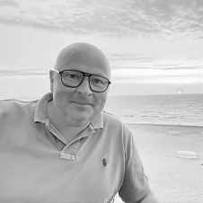 Een persoon met een bril en een poloshirt staat op het strand, met een bewolkte lucht en de oceaan op de achtergrond.