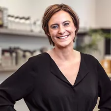 Een glimlachende persoon met kort haar draagt een zwart bloesje en staat binnen met zijn handen in zijn zij. De achtergrond is wazig met planken en potten.