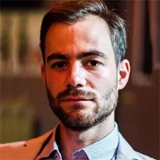 Een man met kort haar en een baard draagt een lichtgekleurd jasje en een geruit overhemd en kijkt in de camera.