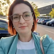 Een persoon met een bril en een blauw jasje maakt een selfie op een parkeerplaats, met auto's en bomen op de achtergrond.