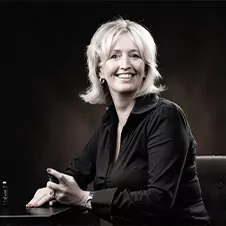 Een vrouw met blond haar zit aan een tafel, glimlachend en met een pen in haar hand tegen een donkere achtergrond. Ze draagt een donker shirt met lange mouwen.