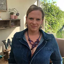 Een persoon draagt een spijkerjack en paarse sieraden en staat binnen, met planten en decoratieve voorwerpen op de achtergrond.