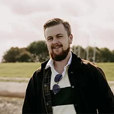 Een man met een baard staat buiten, draagt een jas over een gestreept shirt met een zonnebril die aan de halslijn hangt. Een grasveld en bomen zijn op de achtergrond.