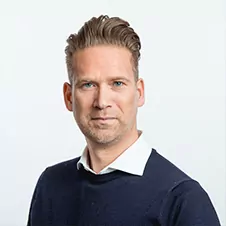 Man met kort, gestyled haar, gekleed in een marineblauwe trui over een wit overhemd, staande tegen een effen, lichte achtergrond.