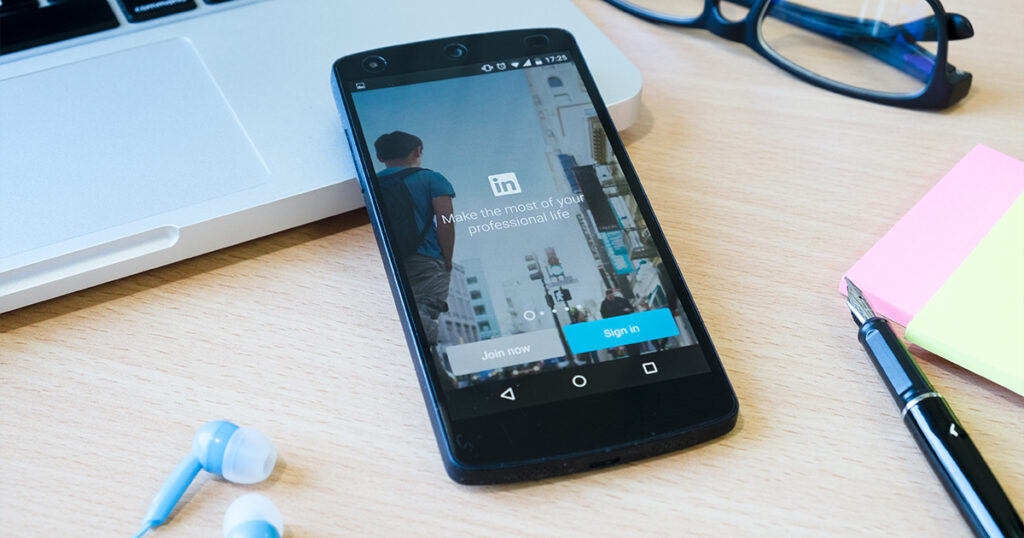 Een smartphone met het LinkedIn-inlogscherm op het bureau, naast een laptop, bril, oordopjes, plakbriefjes en een pen. Dit legt de essentie van netwerken vast in het tijdperk van algoritmes en virale content.