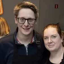 Twee mensen staan dicht bij elkaar binnenshuis en lachen allebei naar de camera.