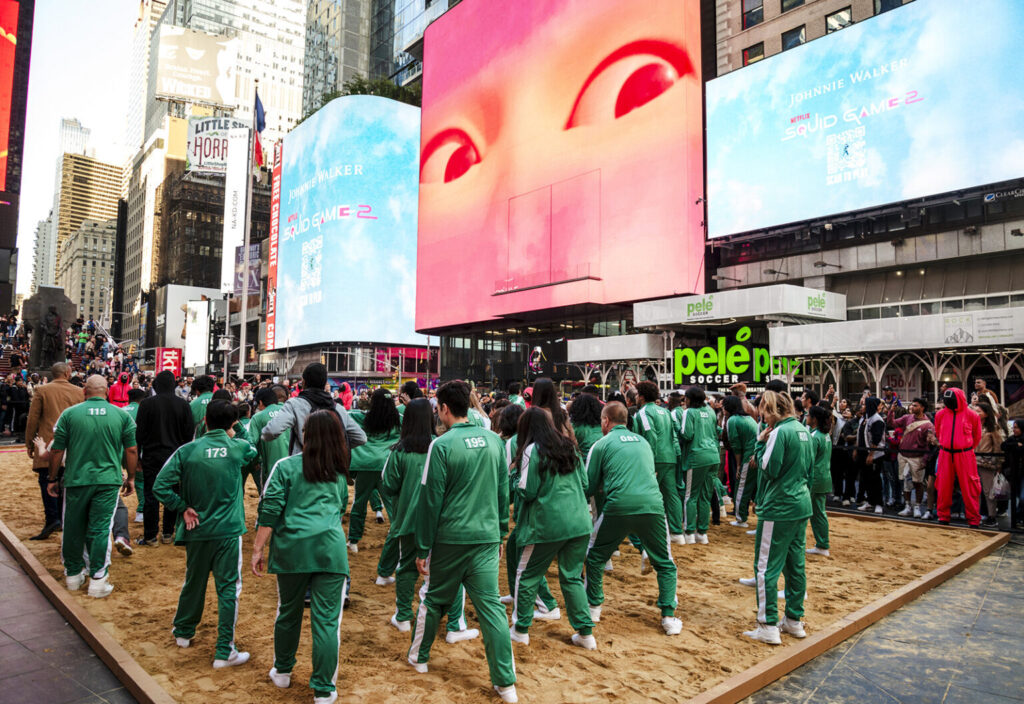 Een groep mensen in groene trainingspakken verzamelt zich op een zanderig gebied op een stadsplein, omringd door grote elektronische billboards. Het lijkt wel een live Netflix-marketingstrategie.