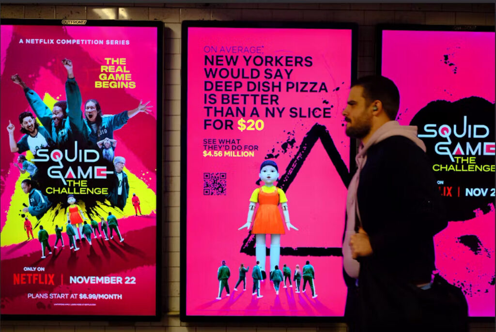Iemand loopt langs drie verlichte posters voor 'Squid Game: The Challenge'. Deze posters laten de marketingstrategie van Netflix zien met opvallende afbeeldingen en teksten die de release van de serie op een opvallende manier promoten. Tegelijkertijd is het een hot topic op sociale-mediaplatforms.