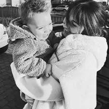 Een vrouw houdt een jong kind buiten in Limburg vast en lacht ernaar. Beiden zijn warm gekleed en lijken gelukkig. Het beeld, in zwart-wit, legt de vreugde van het moment vast.