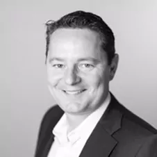 Zwart-wit kopfoto van een man met een colbert en overhemd met kraag, lachend naar de camera tegen een effen achtergrond.