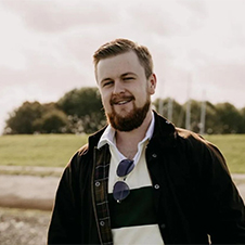 Een man met een baard staat buiten, draagt een jas over een gestreept shirt met een zonnebril die aan de halslijn hangt. Een grasveld en bomen zijn op de achtergrond.