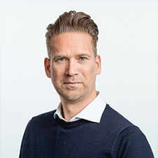 Man met kort, gestyled haar, gekleed in een marineblauwe trui over een wit overhemd, staande tegen een effen, lichte achtergrond.